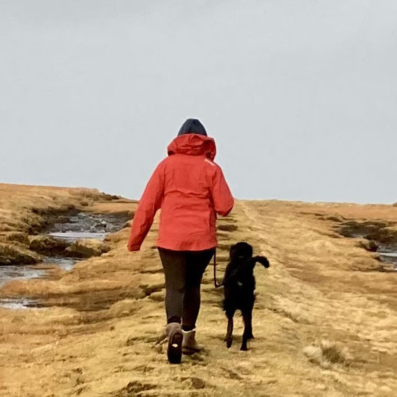 Mey in einer roten Jacke und schwarzer Hose mit braunen Stiefeln von hinten. Rechts neben Mey läuft ein schwarzer Hund. Die beiden gehen auf hellgelbem Gras, der Himmel ist hell graublau.